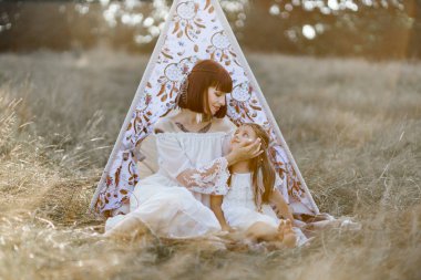 Anne küçük kızının yüzüne hafifçe dokunur, yaz tarlasında çadırın yanında birlikte oturur.