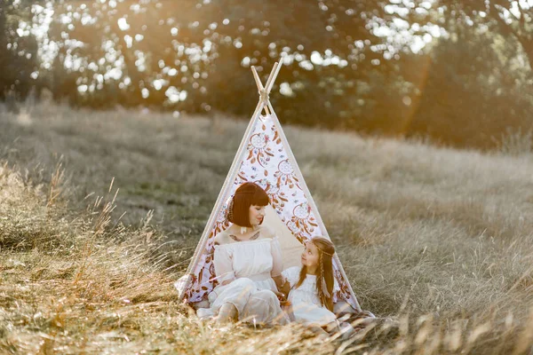 Maman et petite fille portant une robe blanche, accessoires de coiffure de plumes boho et assis près de la tente de tipi — Photo