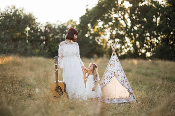 Жінка ходить з гітарою на полі на заході сонця, разом зі своєю чарівною маленькою донькою — стокове фото