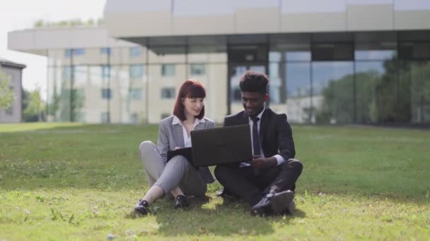 Mutliethnic colegas masculinos y femeninos que trabajan con el ordenador portátil fuera de la oficina sentado en la hierba verde — Vídeos de Stock