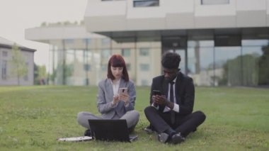 İki çok ırklı iş arkadaşı, çimenlerin üzerinde oturup tatilin tadını çıkarıyor, telefonları kullanıyor ve beğenilerini gösteriyor.