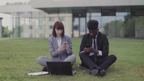 Compañeros de negocios multiétnicos, disfrutando de un descanso al aire libre, sentados en la hierba cerca de la oficina, usando teléfonos — Vídeo de stock