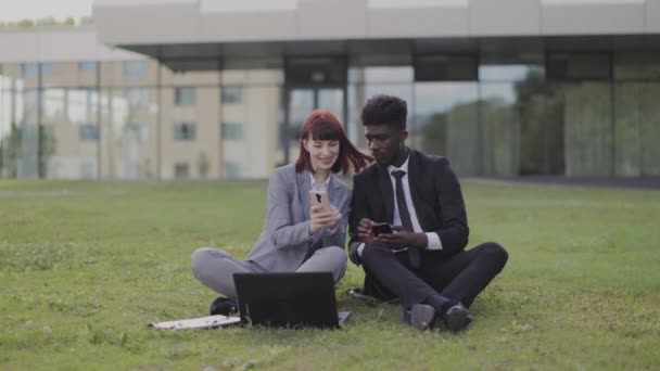 Geschäftsfrau sitzt draußen auf Gras und zeigt ihrem afroamerikanischen Kollegen etwas am Telefon — Stockvideo