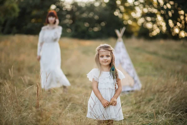 白いドレスと髪の毛の羽のかなり小さな女の子,日没で夏のフィールドのカメラでポーズ. — ストック写真