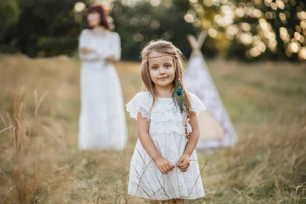 Menina adorável em vestido e penas, de pé no campo de verão, enquanto sua mãe bonita fica atrás — Fotografia de Stock