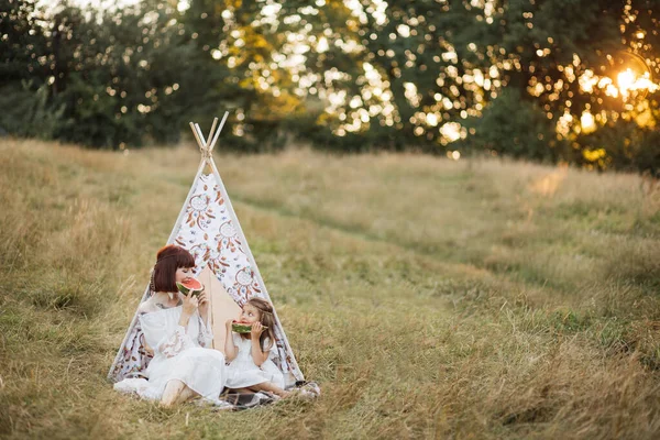 Mãe e sua linda garota, sentado perto tee xixi barraca no campo e mordendo melancia fresca — Fotografia de Stock