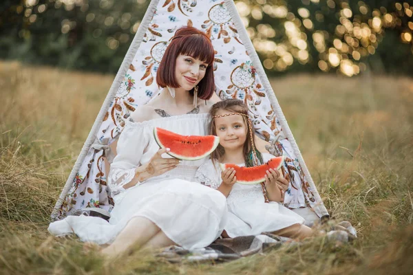 Mãe e filha feliz, sentado na frente de boho wigwam teepee com pedaços de melancia — Fotografia de Stock