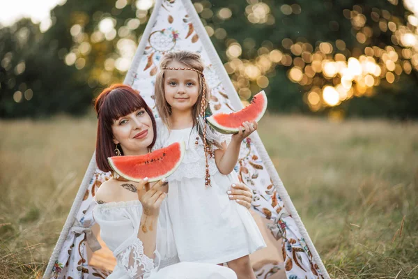 Mãe e filha bonito vestido em estilo boho, sentado na frente da tenda tee-pee com melancia — Fotografia de Stock