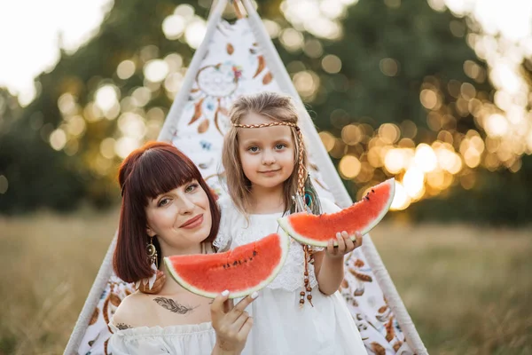 Muito boho hippie mãe com filha sentada perto da tenda wigwam e comer melancia — Fotografia de Stock