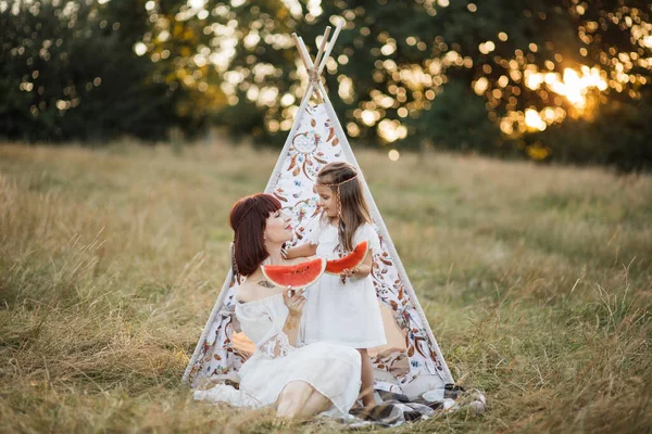 Mãe e filha, passando seu tempo de piquenique no campo, sentados perto de wigwam com melancia — Fotografia de Stock