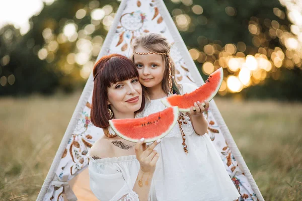 Muito boho hippie mãe com filha sentada perto da tenda wigwam e comer melancia — Fotografia de Stock