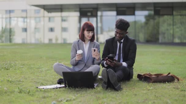 Compañeros sonrientes, sentados en la hierba al aire libre y usando el teléfono celular, tomando café — Vídeos de Stock