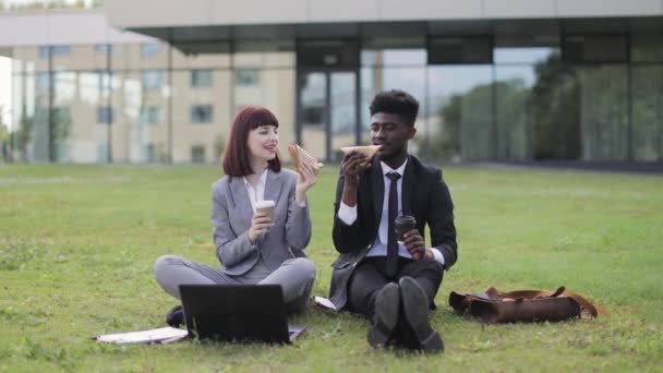 Collègues d'affaires multiethniques, assis sur l'herbe à l'extérieur du bureau, manger des sandwichs pendant le déjeuner — Video