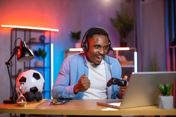 Homme africain dans un casque célébrant la victoire dans le jeu vidéo sur ordinateur portable avec les poings serrés et sourire — Photo