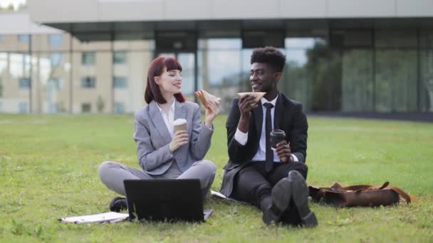 Deux jeunes gens d'affaires qui déjeunent ensemble, assis sur l'herbe à l'extérieur — Video