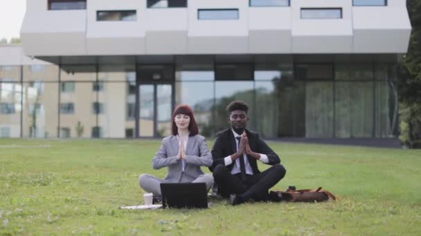 Двоє багатоетнічних бізнесменів, сидячи на зеленій траві перед офісом, роздумуючи з закритими очима — стокове відео