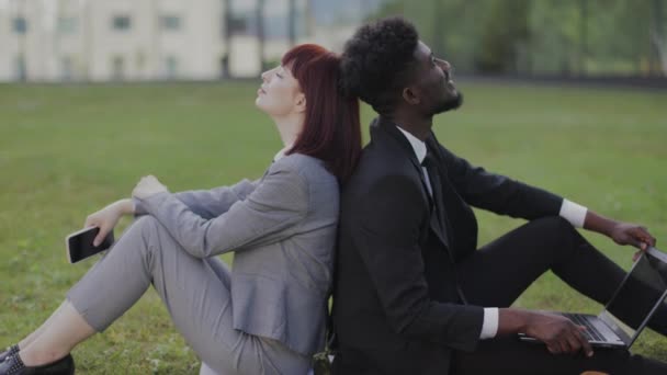 Multiethnique deux collègues de bureau d'affaires, assis dos à dos sur l'herbe et se détendre pendant la pause — Video