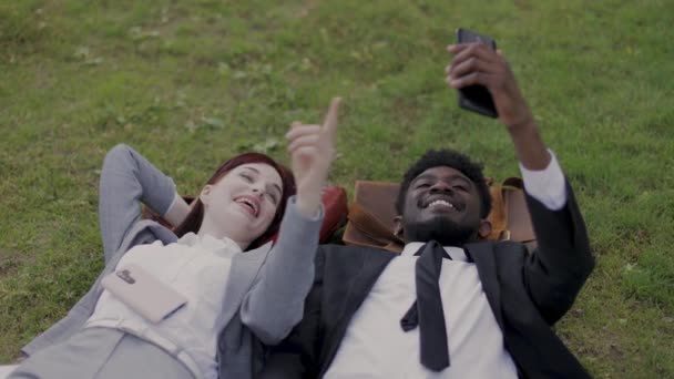 Çok ırklı, iyi giyimli genç iş adamları akıllı telefonlu yeşil çimenlerin üzerinde dinleniyorlar. — Stok video