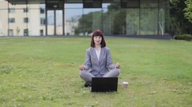 Sakin genç bir kadın meditasyon yapıyor, dışarıda yeşil çimlerde dizüstü bilgisayarla oturuyor.