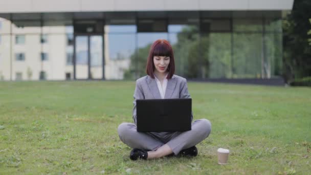 Mujer bonita en traje de negocios, sentada con piernas cruzadas sobre hierba verde usando una computadora portátil, celebrando el éxito — Vídeo de stock