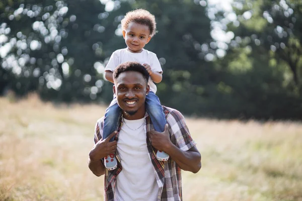 Africano niño sentado en padre hombros durante paseo al aire libre — Foto de Stock