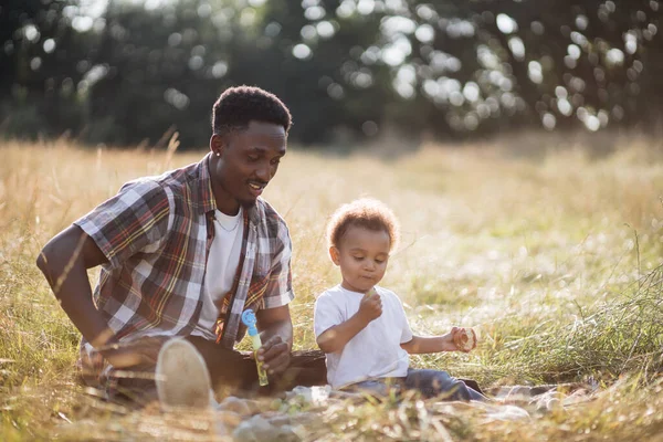 Pai e filho africanos brincando com bolhas de sabão ao ar livre — Fotografia de Stock