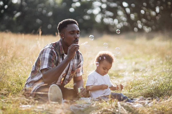 Africano homem e bonito menino brincando com bolhas de sabão no campo — Fotografia de Stock