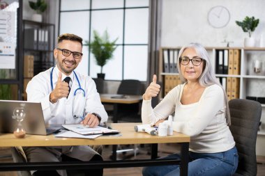 Doktor ve hasta kamerada baş parmağını gösteriyor ve gülümsüyor.