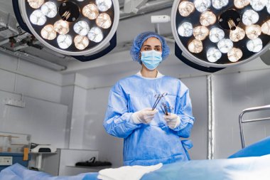 A female surgeon stands in an operating room, holding surgical tools and wearing a mask and scrubs.