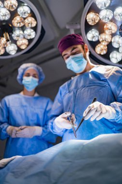 A surgeon and assistant are performing an operation in a modern operating room, illuminated by bright surgical lights.