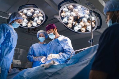 A surgical team performs an operation in a modern operating room, illuminated by bright overhead lights.