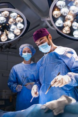 A surgeon and assistant perform surgery in a modern operating room, illuminated by bright surgical lights.
