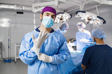 A surgeon wearing a mask and gloves in an operating room, preparing for a procedure with his team in the background.