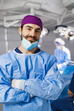 A surgeon, wearing a surgical mask and scrubs, stands in an operating room with arms crossed, looking at the camera.