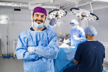 A smiling surgeon in scrubs stands in an operating room with his arms crossed, with other medical professionals in the background.