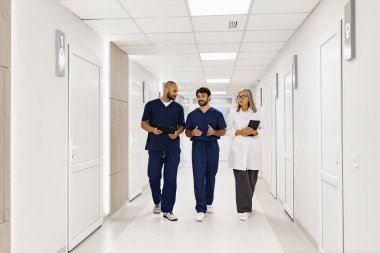 Aralarında doktorlar ve hemşirelerin de yer aldığı bir tıp uzmanları ekibi, muhtemelen vizitler sırasında veya danışmanlık sırasında, parlak bir hastane koridorunda yürüyüp tartışıyor ve tartışıyorlar..