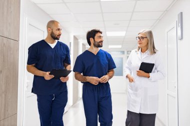 Bir doktor ve iki hemşire hastane koridorunda yürür ve hasta bakımı ve tıbbi prosedürleri tartışırlar..