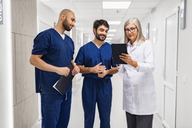 Bir doktor ve iki hemşire klinik koridorunda bir tableti inceleyip hasta bilgilerini ve tıbbi kayıtları tartışıyorlar..