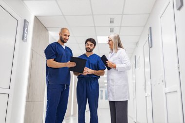 Üç tıp uzmanı, parlak bir hastane koridorunda hasta dosyalarını inceleyip tedavi planlarını tartışıyorlar..