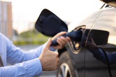 Elektrikli bir aracı şarj ederken, sürdürülebilir taşımacılığa karşı olumlu duygular gösteren bir kişi başparmağını kaldırır.