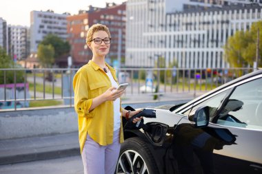 Bir kadın, şehir ortamında elektrikli aracını şarj ederken gülümsüyor, telefonunu tutuyor..