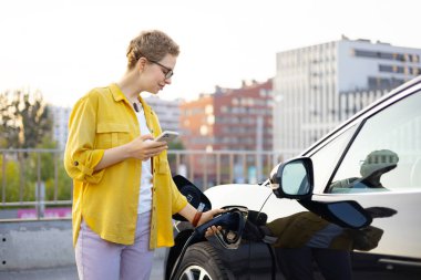 Bir kadın şehir merkezinde elektrikli aracını şarj ederken telefonunu kullanıyor..