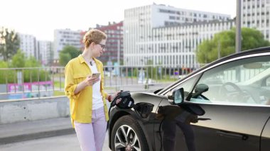 Bir kadın şehir merkezinde elektrikli aracını şarj ederken telefonunu kullanıyor..