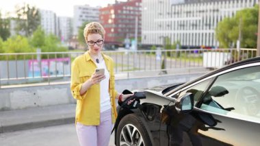 Gülümseyen bir kadın, şehir merkezinde elektrikli aracını şarj ederken telefonunu tutuyor..