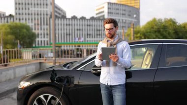 Bir adam elektrikli arabasının yanında durur, tableti tutar ve uzaklığa bakar..