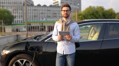 Gözlüklü, gülümseyen bir adam elektrikli arabasının yanında durur ve tableti tutar..