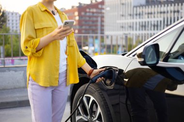 Bir kadın şehir merkezinde elektrikli aracını şarj ederken akıllı telefonunu kullanıyor..