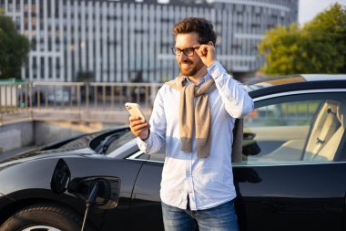 Elektrikli aracı şarj istasyonunda şarj olurken bir adam telefonunu kontrol ediyor..