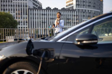 Elinde kahve ve telefon olan bir adam şehir merkezinde şarj olurken elektrikli arabasının yanında duruyor..