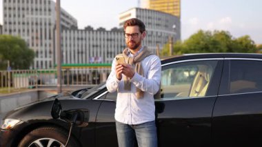 Elektrikli aracı şarj istasyonunda şarj olurken bir adam telefonunu kontrol ediyor..
